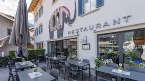 An outdoor seating area with tables and chairs, a large umbrella, and a building with a restaurant sign. - Elefant (Ora)