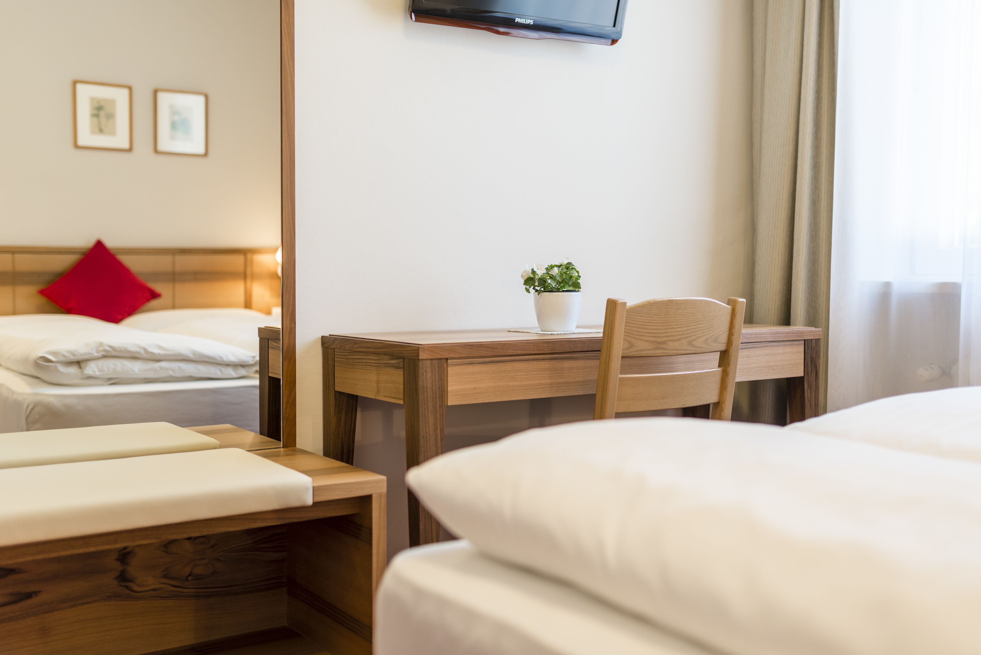 A hotel room with two beds, a wooden desk, a chair, a small potted plant, and a flat-screen TV mounted on the wall.