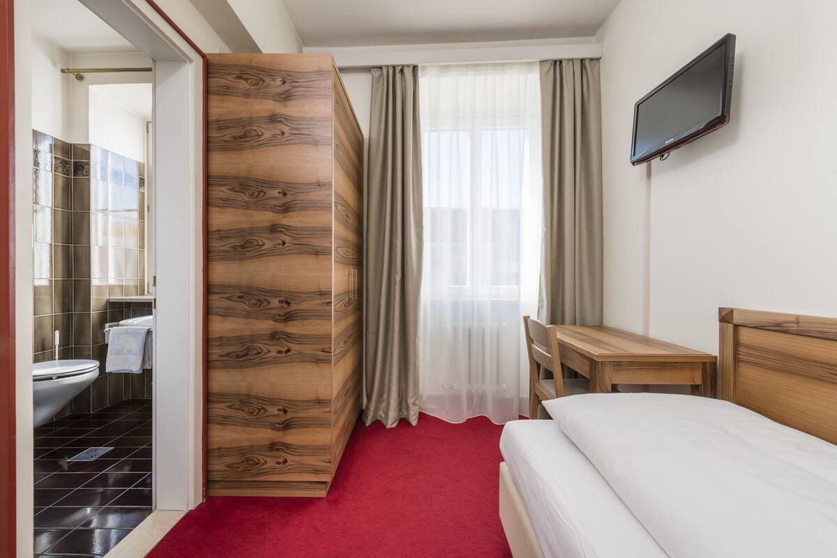 A hotel room with a wooden wardrobe, a bed, a TV mounted on the wall, and a view of the bathroom.