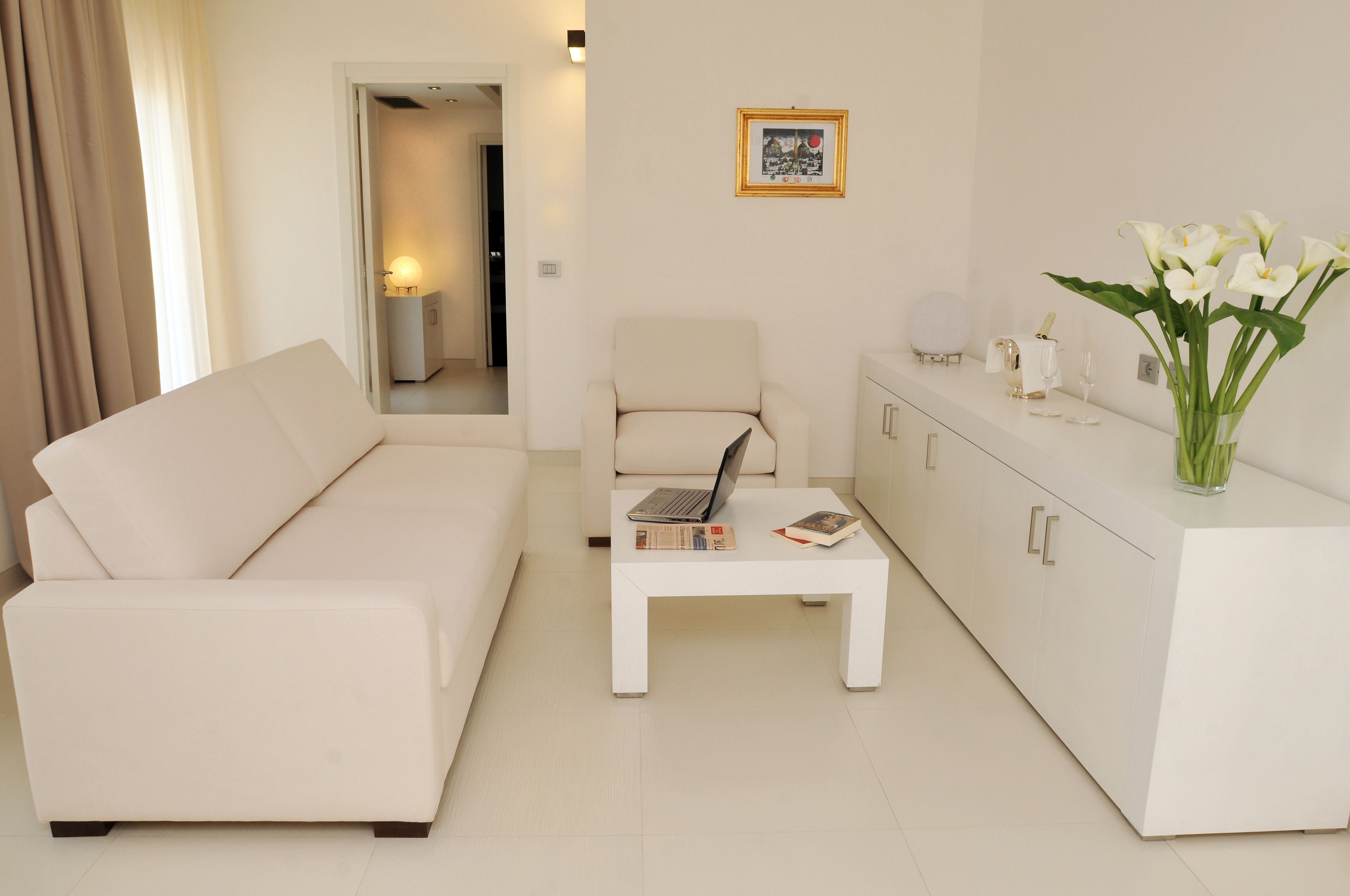 A modern living room with a white sofa, a white coffee table with a laptop and books, a white cabinet with decorative items, and a vase of white flowers.