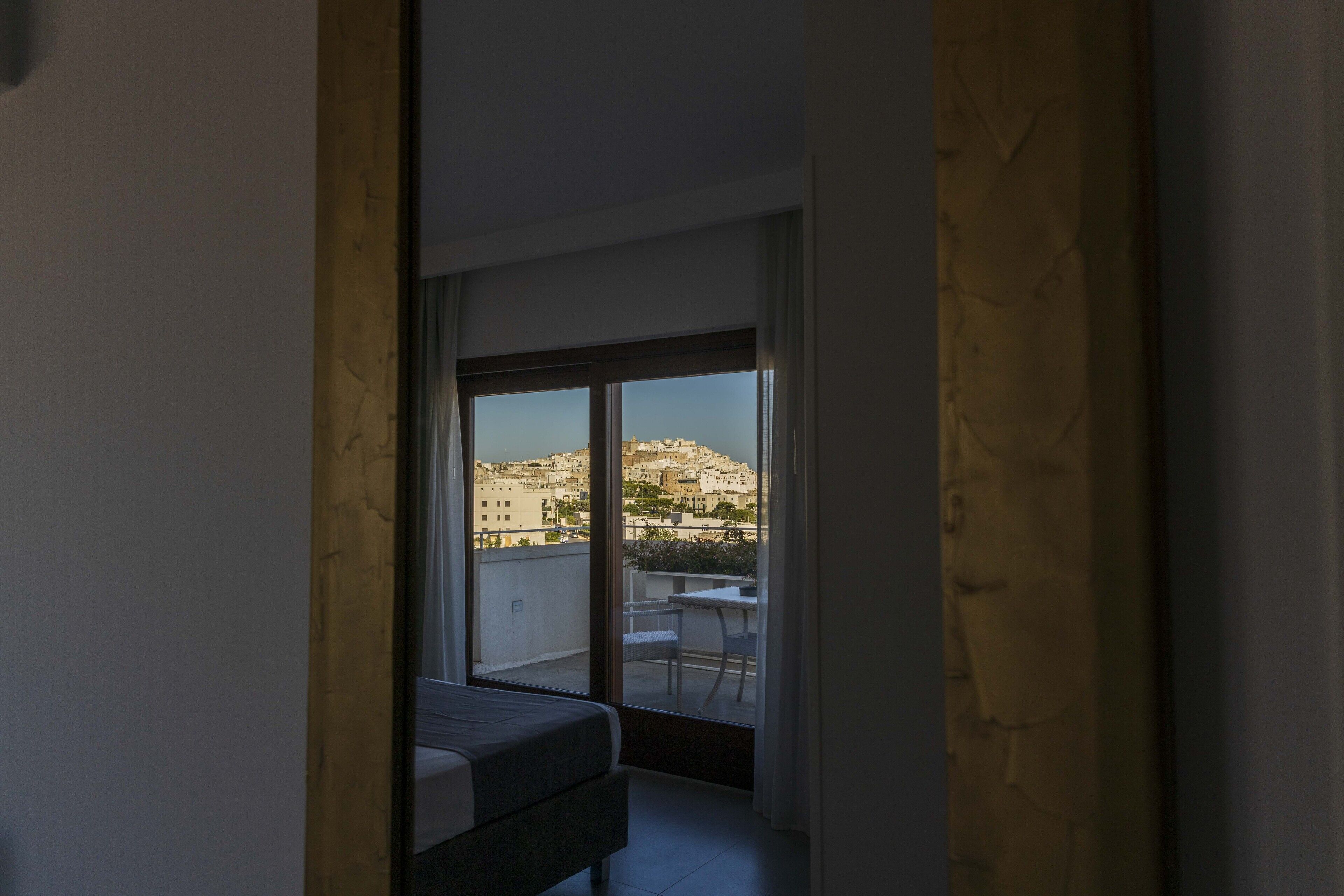 A room with a bed, a small table, and a view of a cityscape through a large window.