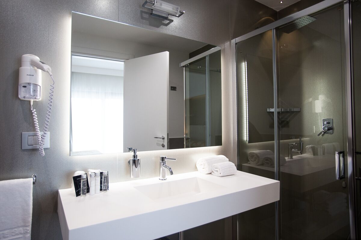 A modern bathroom with a large mirror, a white sink, a hairdryer, and a glass shower enclosure.