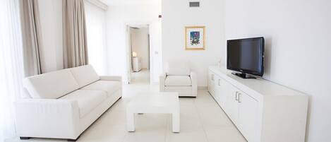 A modern living room with a white sofa, a small coffee table, a white armchair, a TV on a white cabinet, and a framed picture on the wall.
