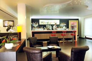 Un bar moderno con un cantinero, taburetes y una variedad de botellas. En primer plano, hay sillas y una mesa, y una planta en maceta sobre la barra.