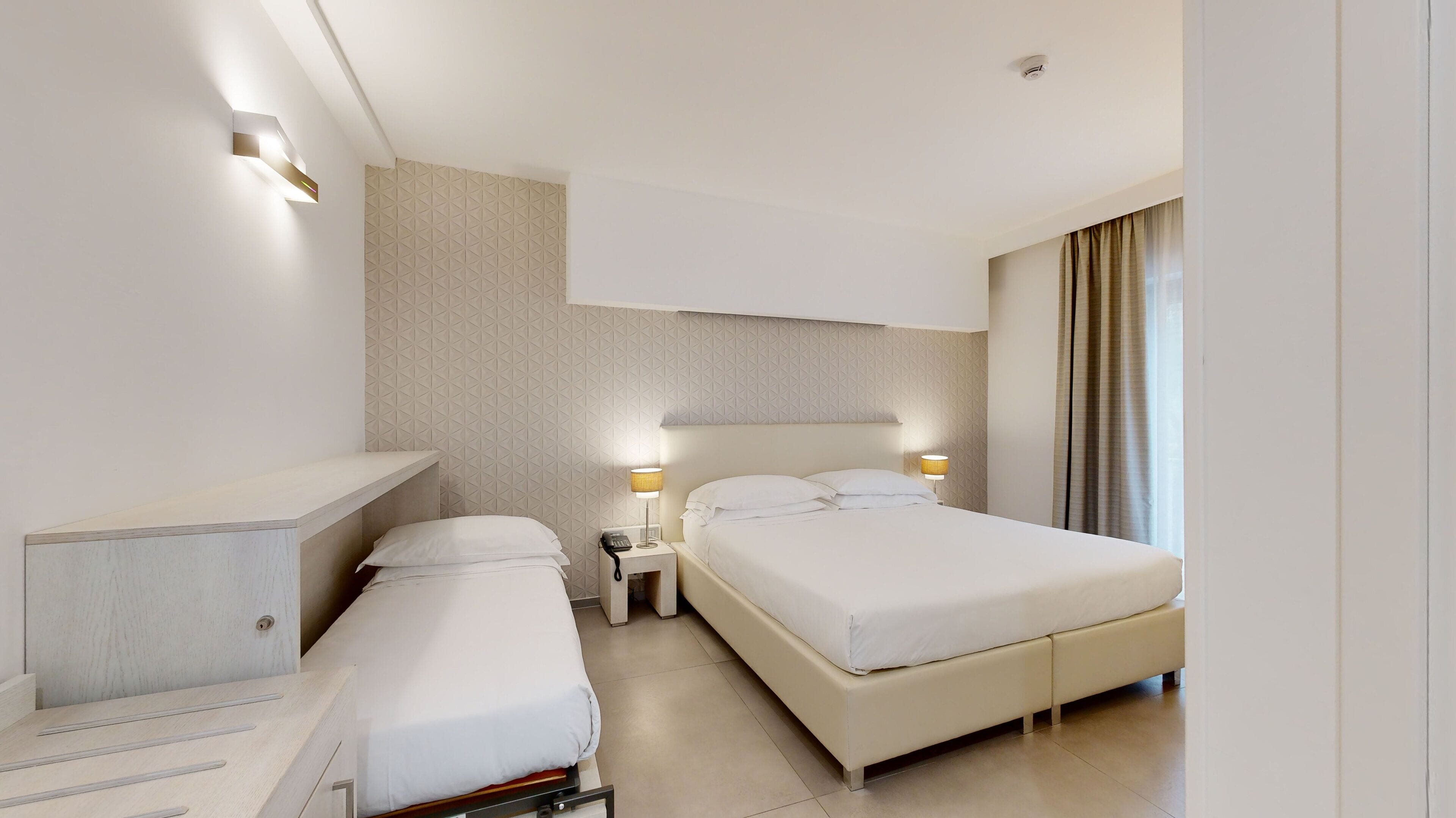 Una habitación de hotel moderna con dos camas individuales, una mesita de noche con lámpara, un estante empotrado y un ventanal con cortinas.