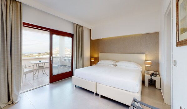 A hotel room with a large bed, a desk with a chair, a sliding glass door to a balcony, and a view of the city.