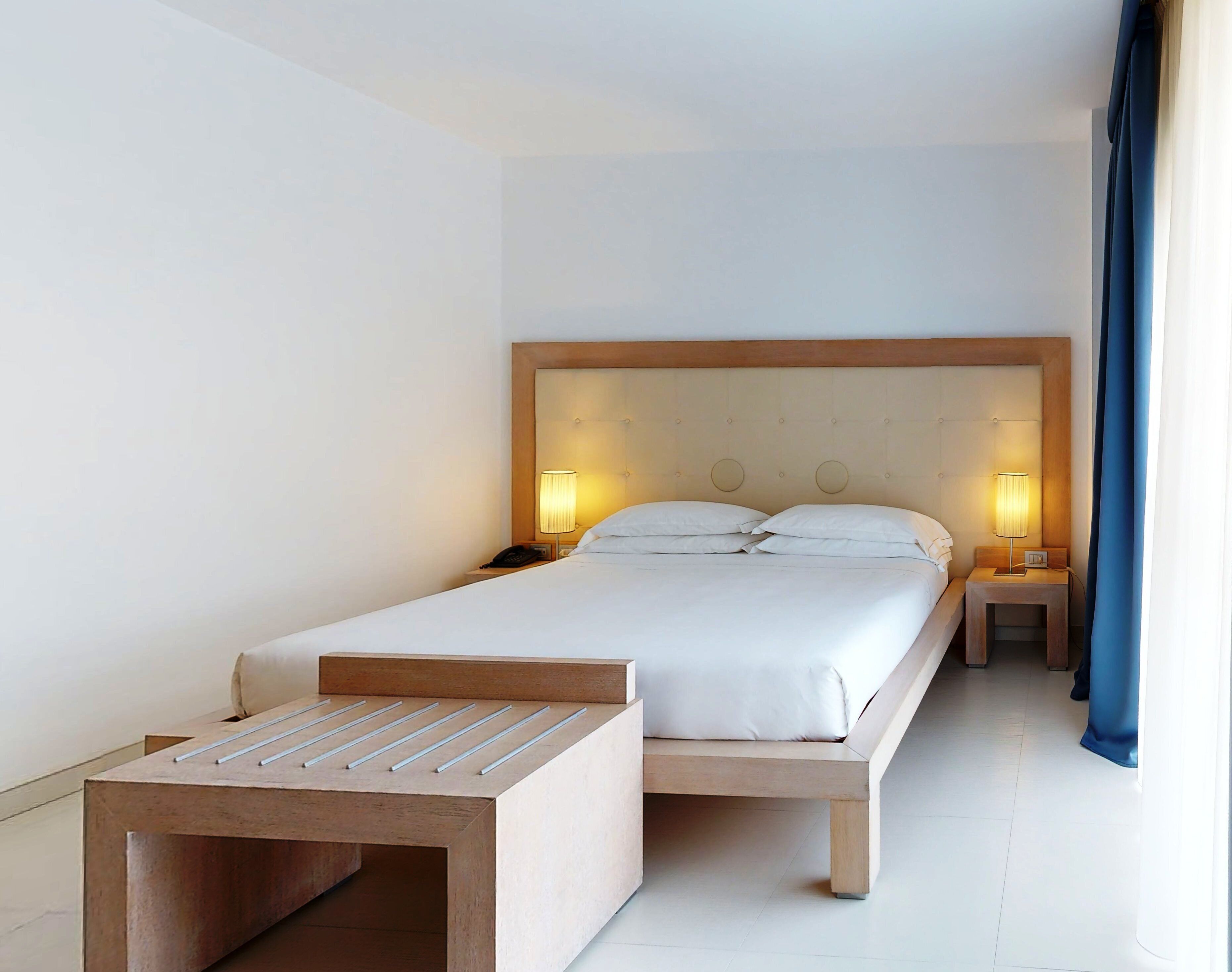 Une chambre d’hôtel moderne avec un grand lit, des lampes de chevet, une table de chevet en bois et une petite table au design à lattes.