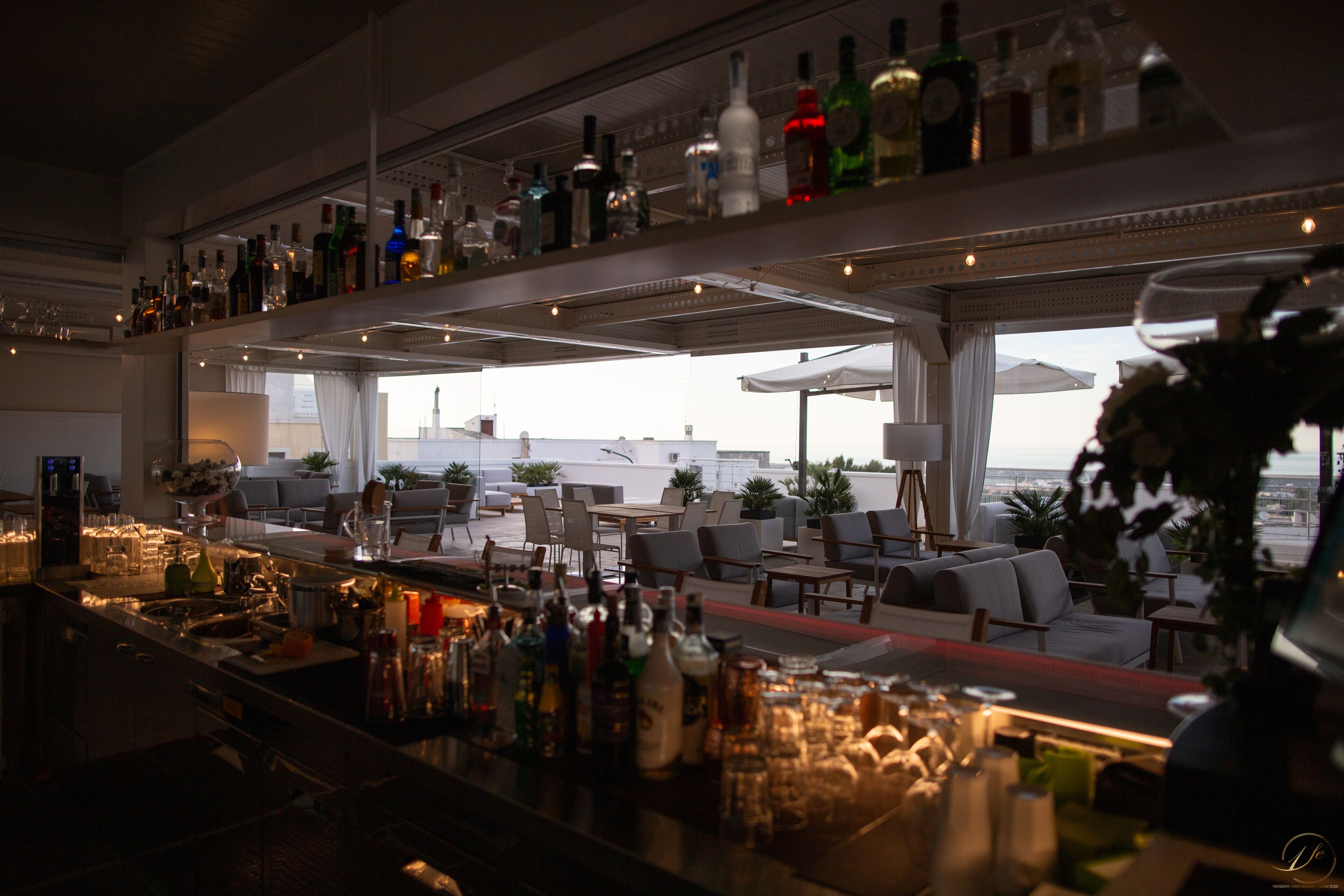 A bar area with a variety of bottles and glasses, a seating area with tables and chairs, and large windows offering a view of the sky.