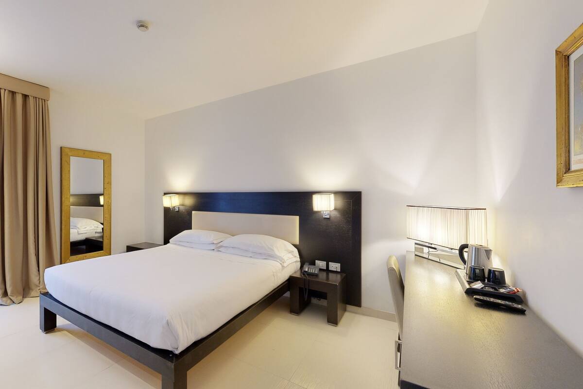 Habitación de hotel con una cama grande, una mesita de noche con lámpara, un espejo y un escritorio con ordenador y teléfono.