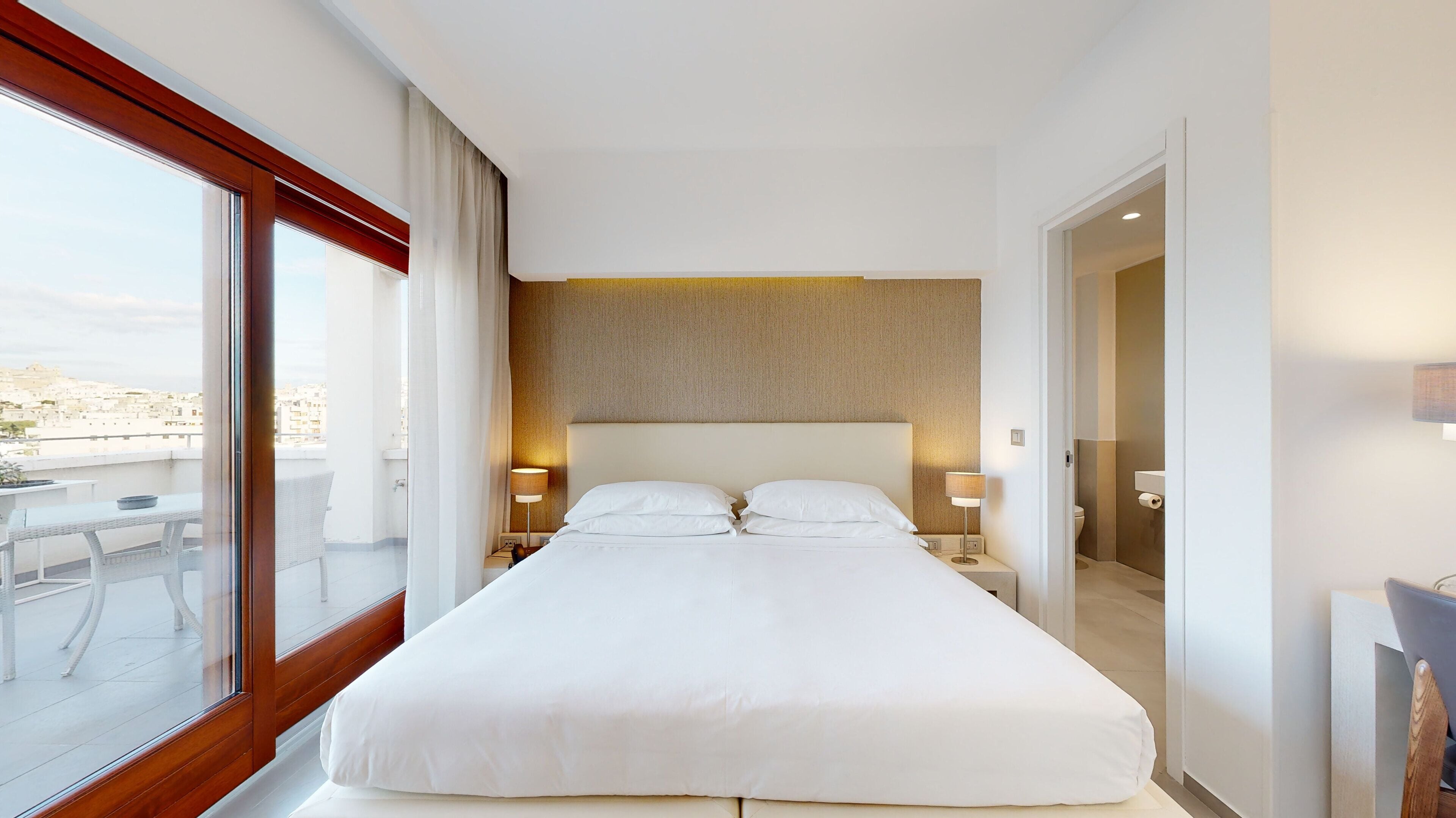 Une chambre d’hôtel avec un grand lit, deux tables de chevet avec des lampes, une vue sur la ville par une grande fenêtre et un balcon avec du mobilier d’extérieur.