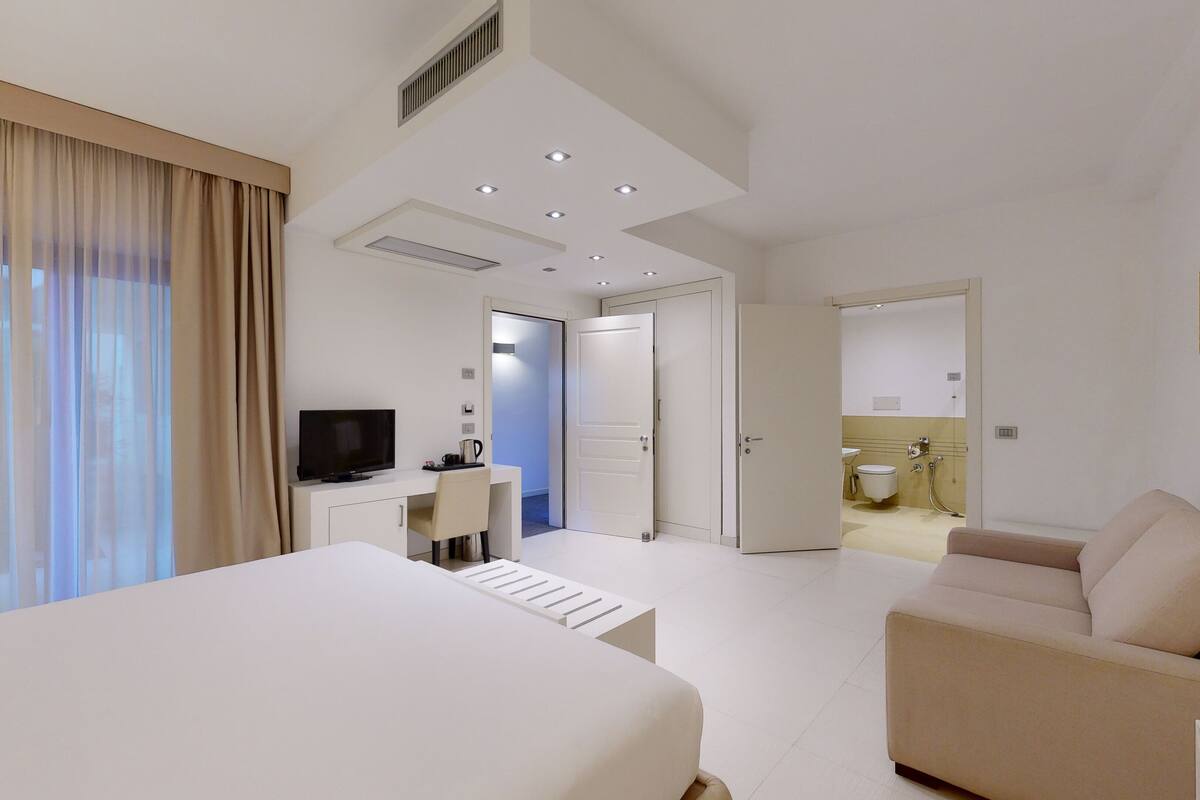 Una habitación de hotel moderna con una cama grande, un escritorio con un televisor de pantalla plana, un sofá y un baño visible a través de una puerta abierta.