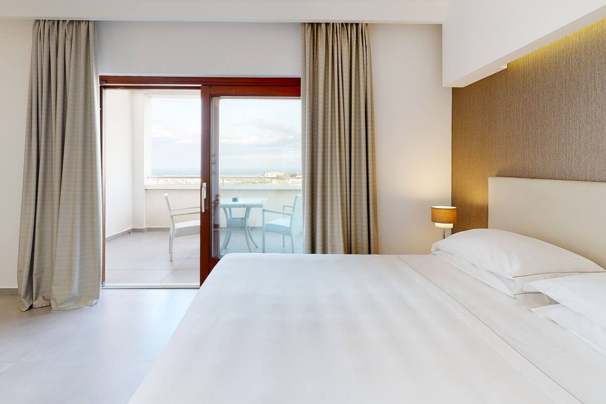 Habitación de hotel con una cama grande, un balcón con mesa y sillas, y vistas a la ciudad.
