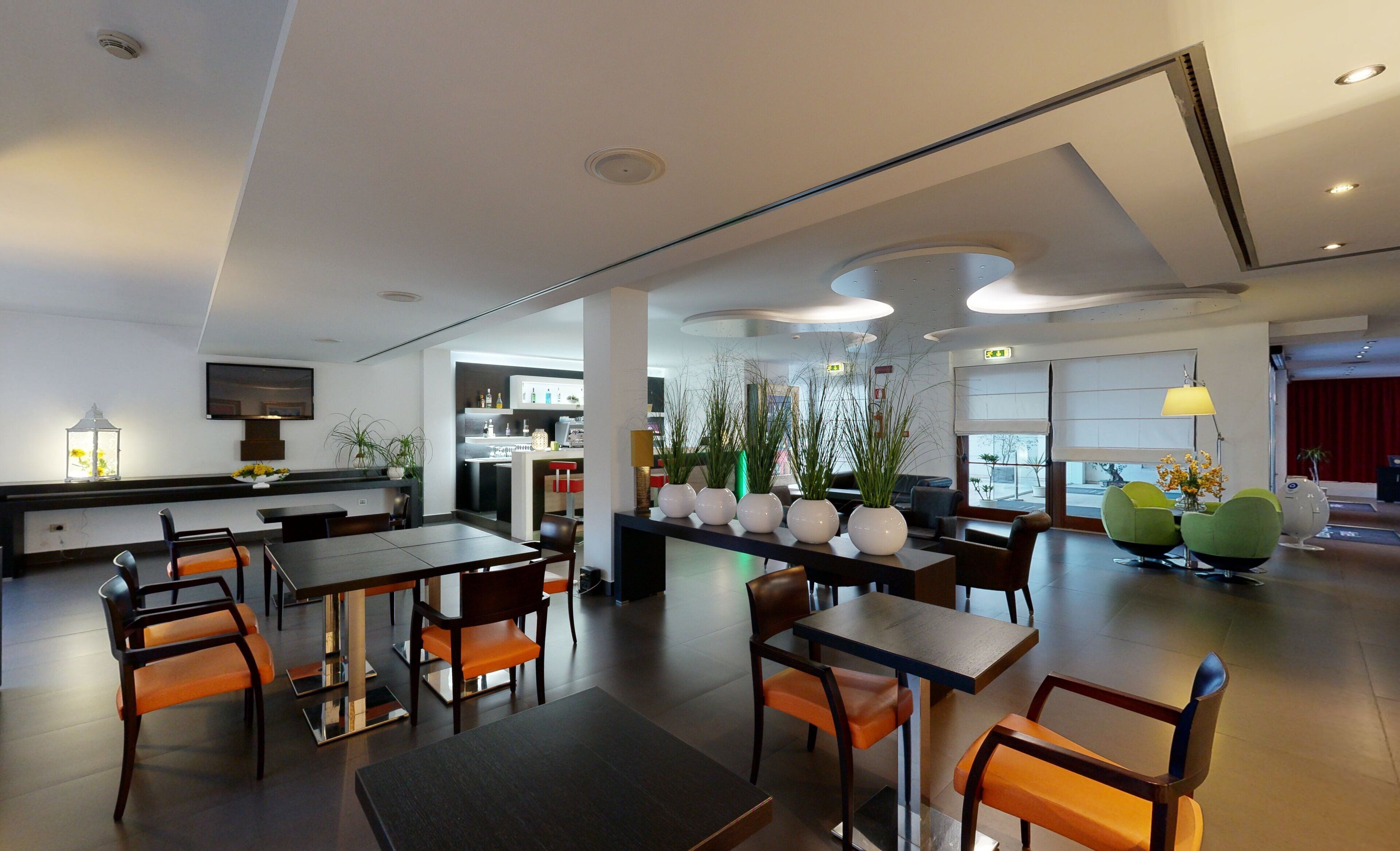 A modern hotel lobby with a dining area, a kitchen in the background, and large windows.