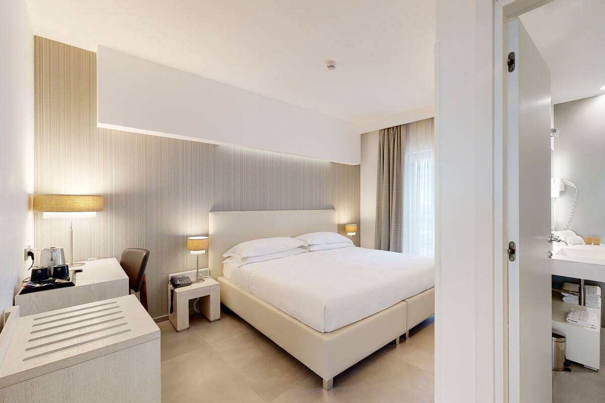 Una habitación de hotel moderna con una cama grande, mesitas de noche con lámparas, un escritorio con lámpara y un baño con lavamanos y espejo.