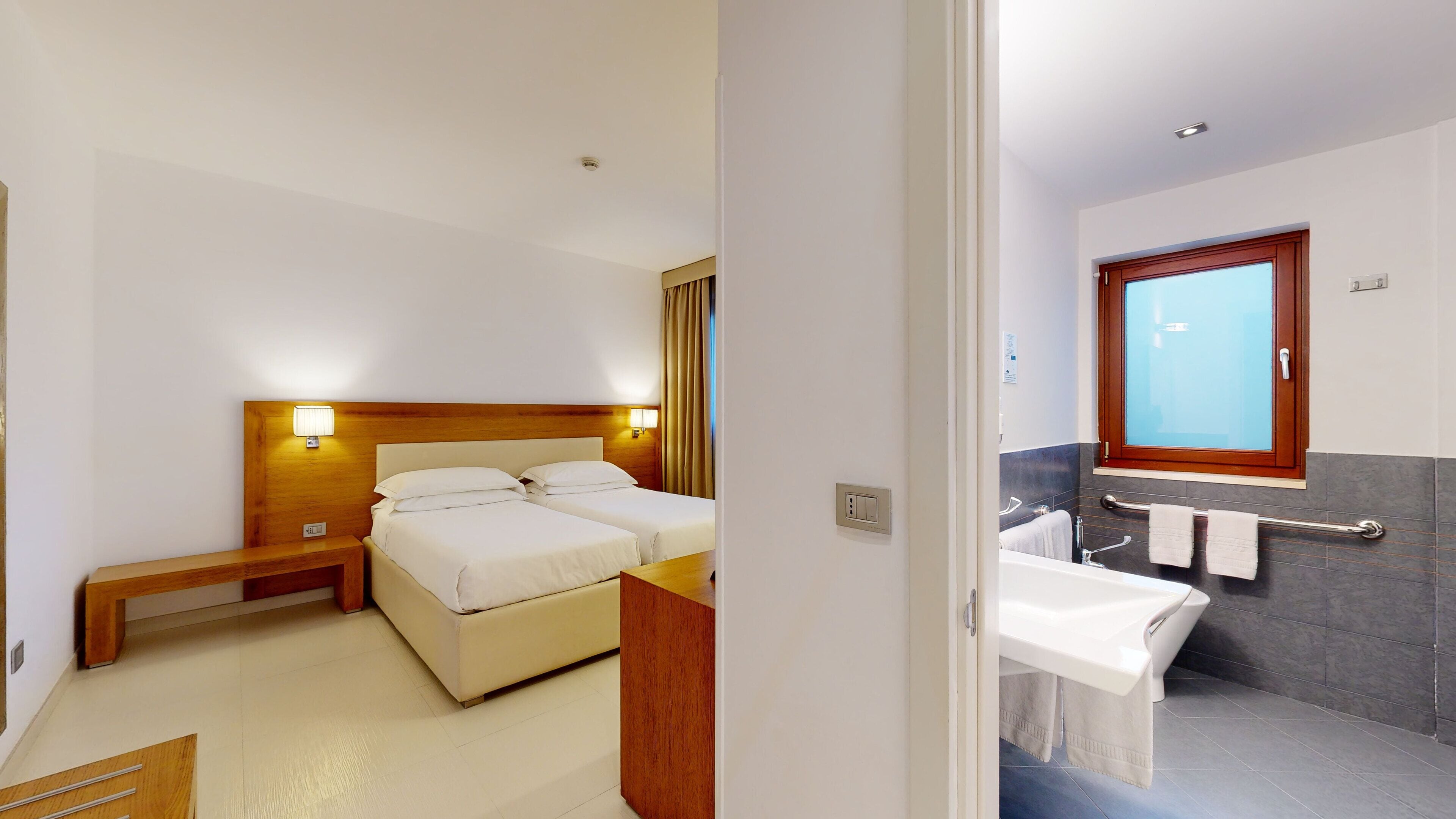 Ett hotellrum med två sängar, en sänggavel i trä, sänglampor, en träbänk och ett badrum med badkar, handfat och handduksställ.