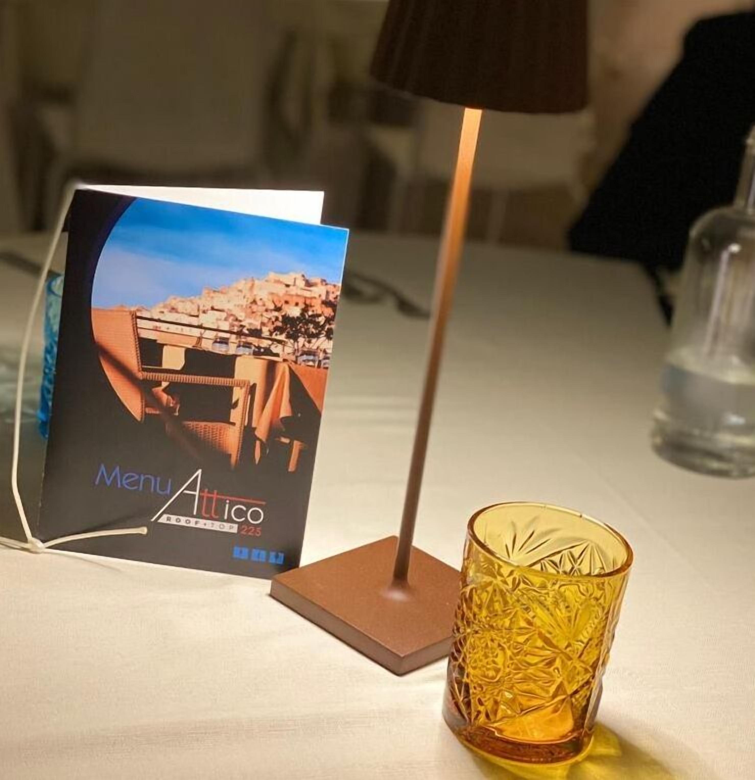 Carta de menú de hotel con una imagen de la ciudad, una lámpara de pie alta y un vaso de vidrio texturizado sobre una mesa.