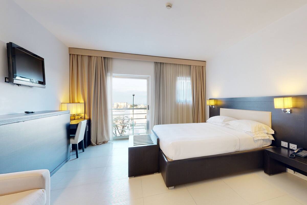 Habitación de hotel con cama, mesita de noche, lámpara, escritorio, silla y televisor en la pared.