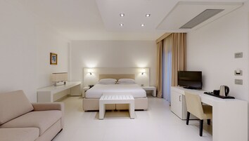 A modern hotel room with a large bed, a desk with a TV, a sofa, and a bedside table with a lamp.