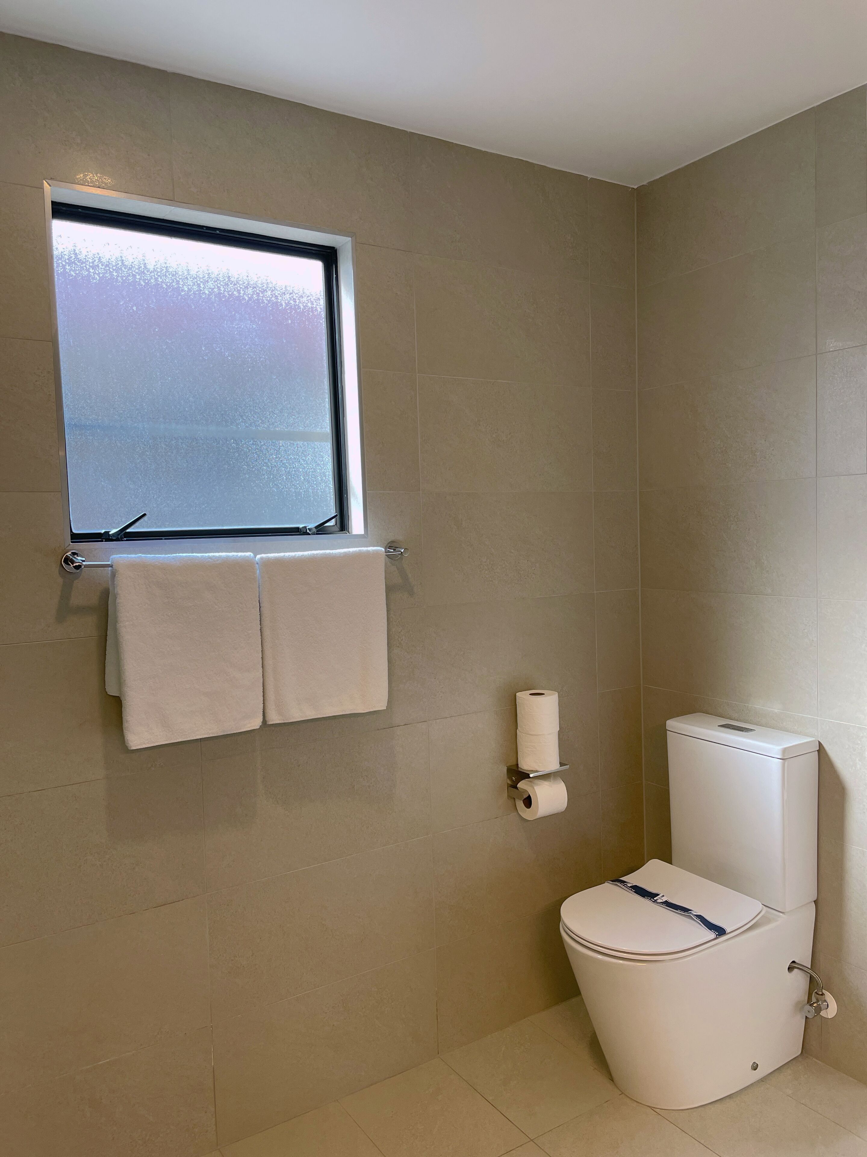 Un baño con inodoro, un perchero con dos toallas, un dispensador de papel higiénico y una ventana con vista al cielo.