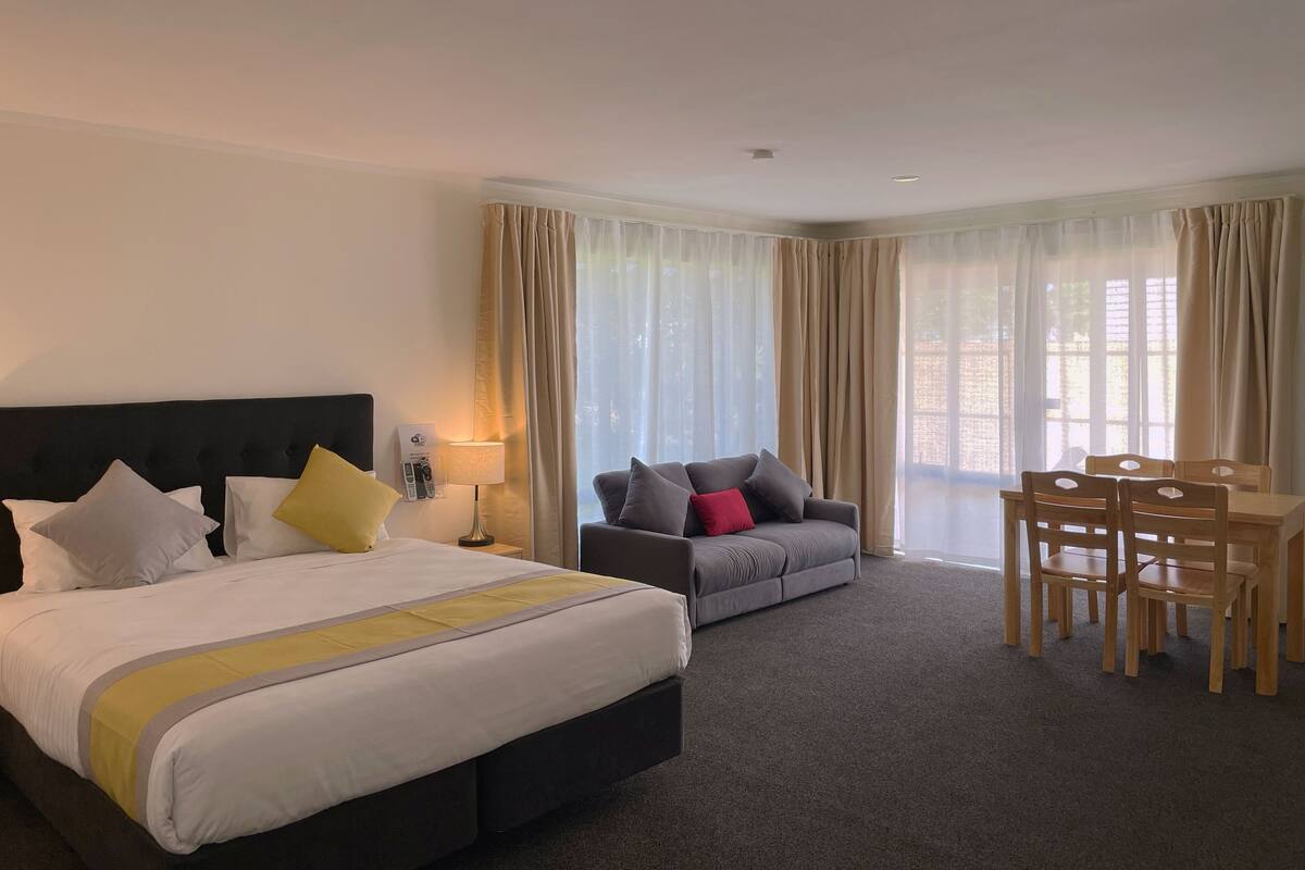 A hotel room with a bed, a sofa, a dining table with chairs, and large windows with curtains.