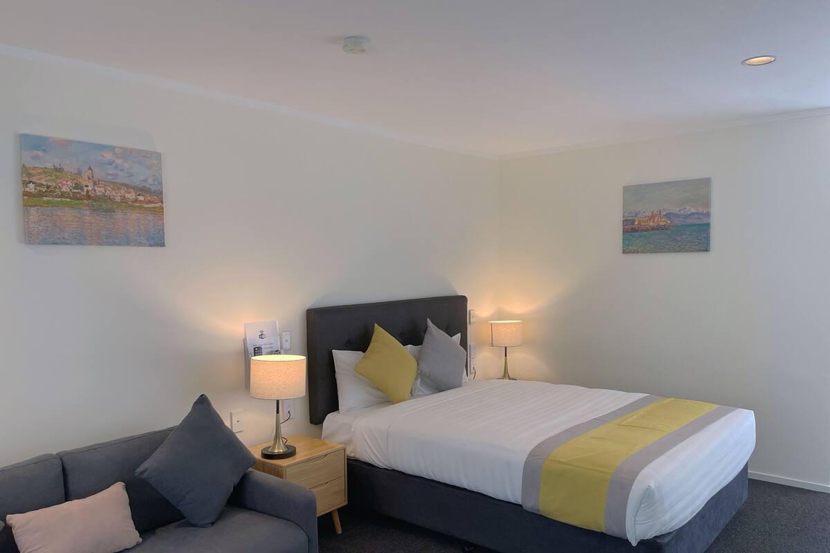 A hotel room with a bed, a bedside table with a lamp, a grey sofa, and two paintings on the walls.