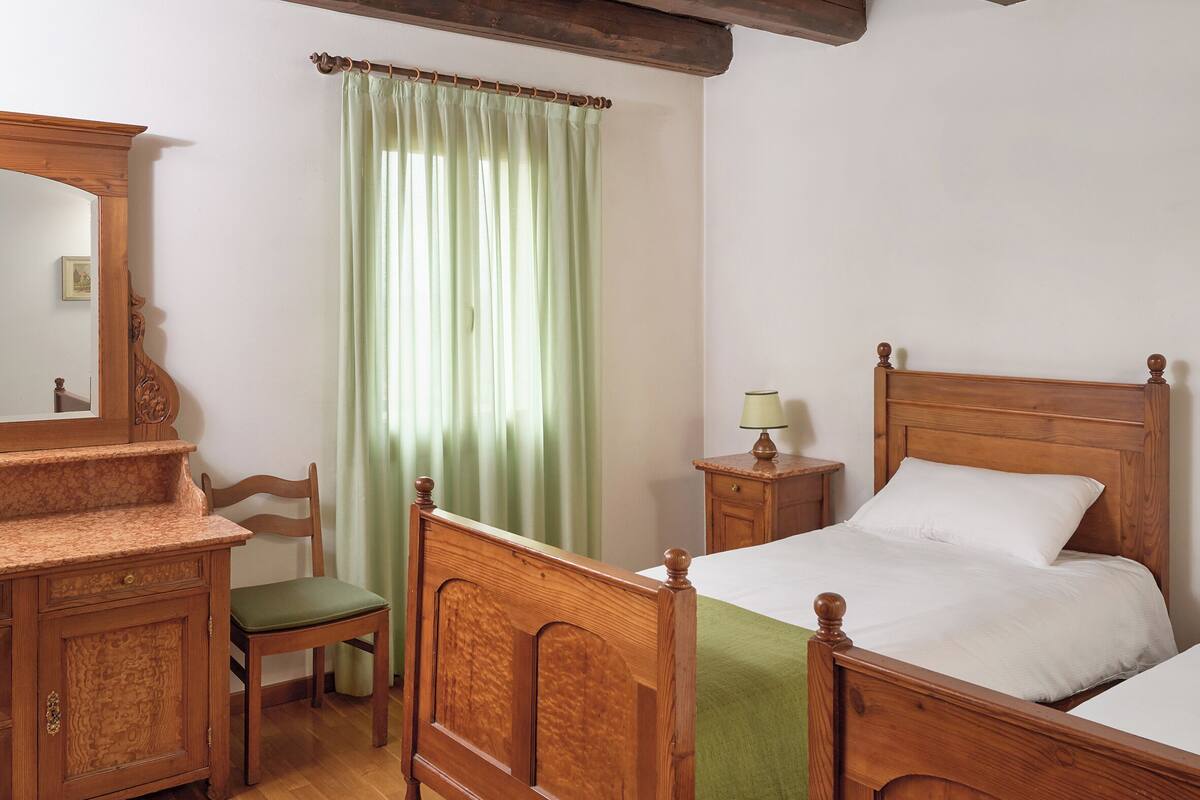 Ein Schlafzimmer mit einem hölzernen Bett, einer Holzkommode, einem Stuhl, einem Nachttisch mit Lampe und einem Fenster mit grünen Vorhängen.