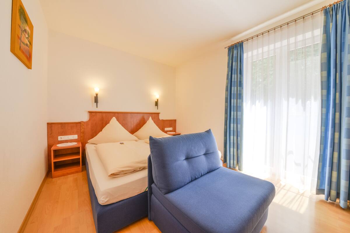 Een hotelkamer met een bed, een blauwe fauteuil, een houten nachtkastje en een raam met dunne gordijnen.