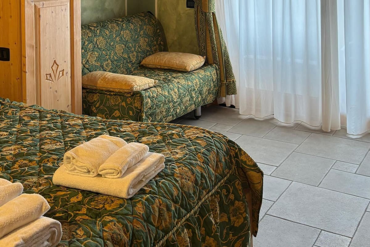 A bedroom with a wooden wardrobe, a bed with a patterned bedspread, a green sofa with cushions, and a window with sheer curtains.