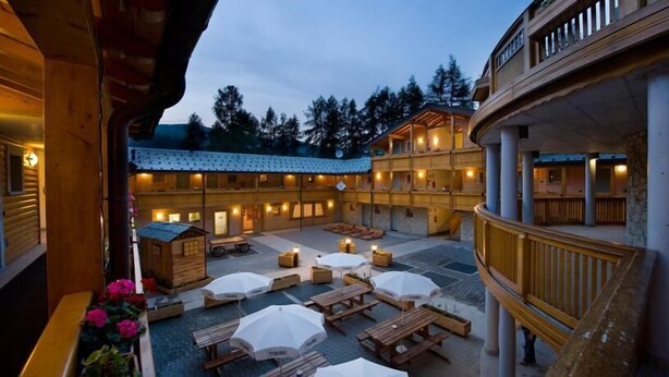 A lodge with multiple wooden buildings, outdoor seating areas with umbrellas, and a view of the surrounding forest. - Boscone Suite Hotel (Madesimo)