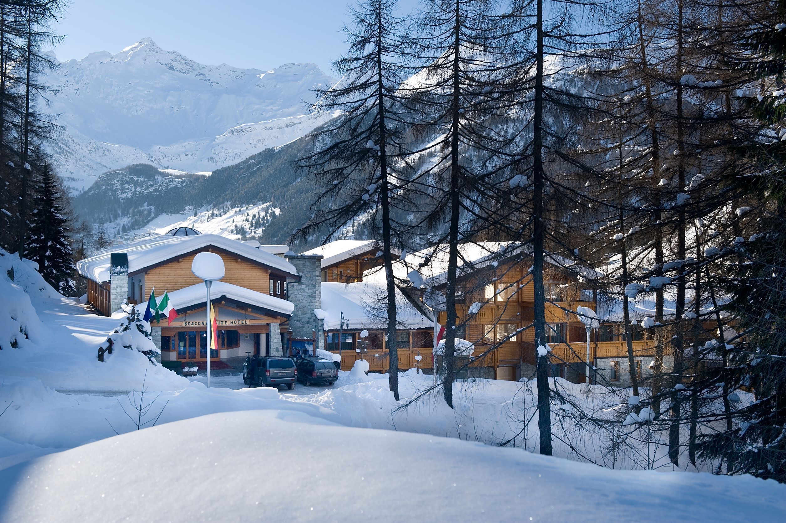 Un chalet enneigé, avec une façade en bois et une entrée en pierre, entouré d’arbres et de montagnes couverts de neige.