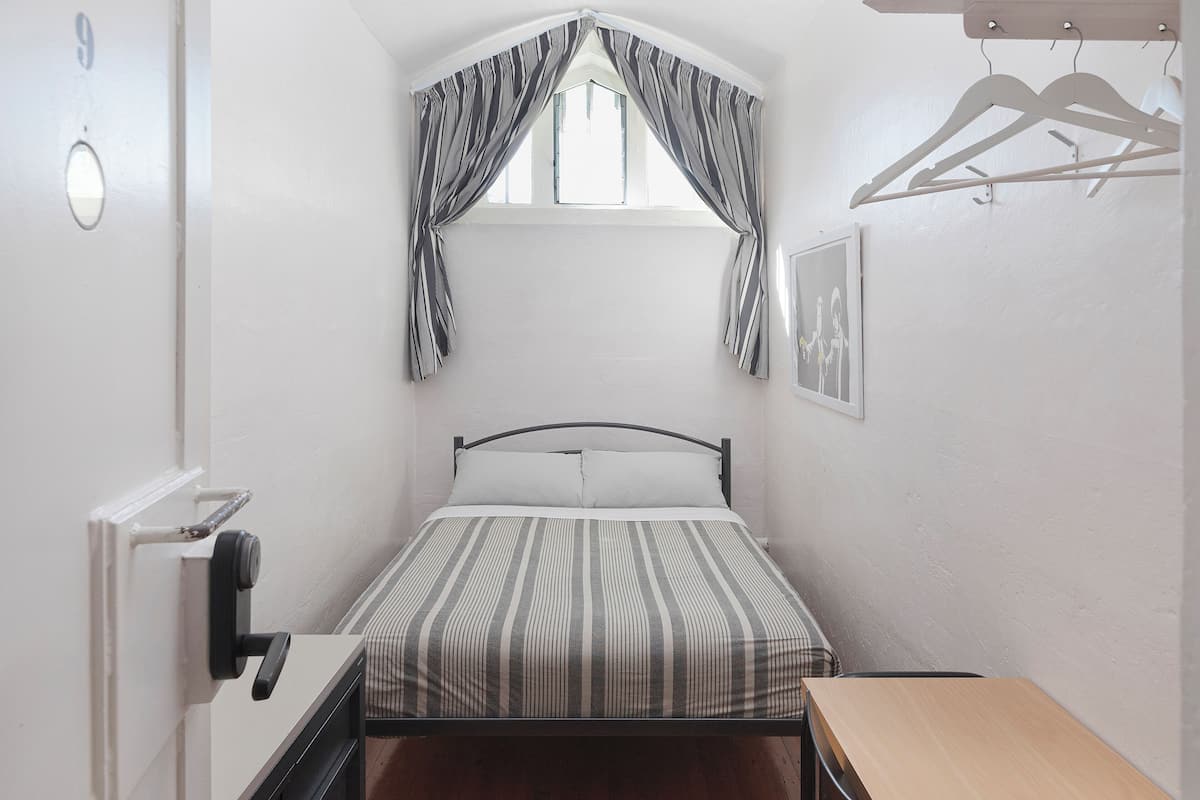Una habitación pequeña con cama individual, ventana, ropa de cama a rayas, un escritorio de madera y un perchero con ganchos.