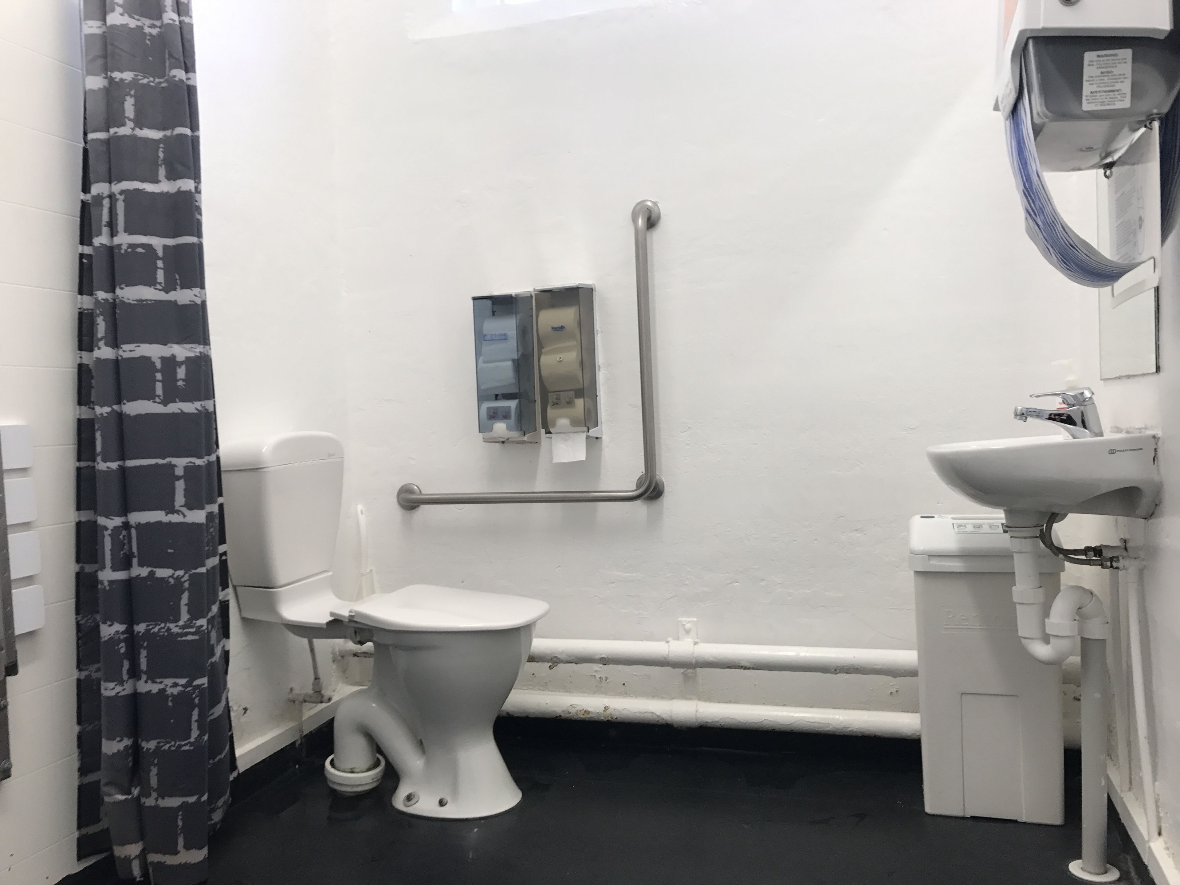 Un baño con inodoro blanco, lavamanos y secamanos. Cuenta con un pasamanos en la pared y una cortina con estampado blanco y negro.