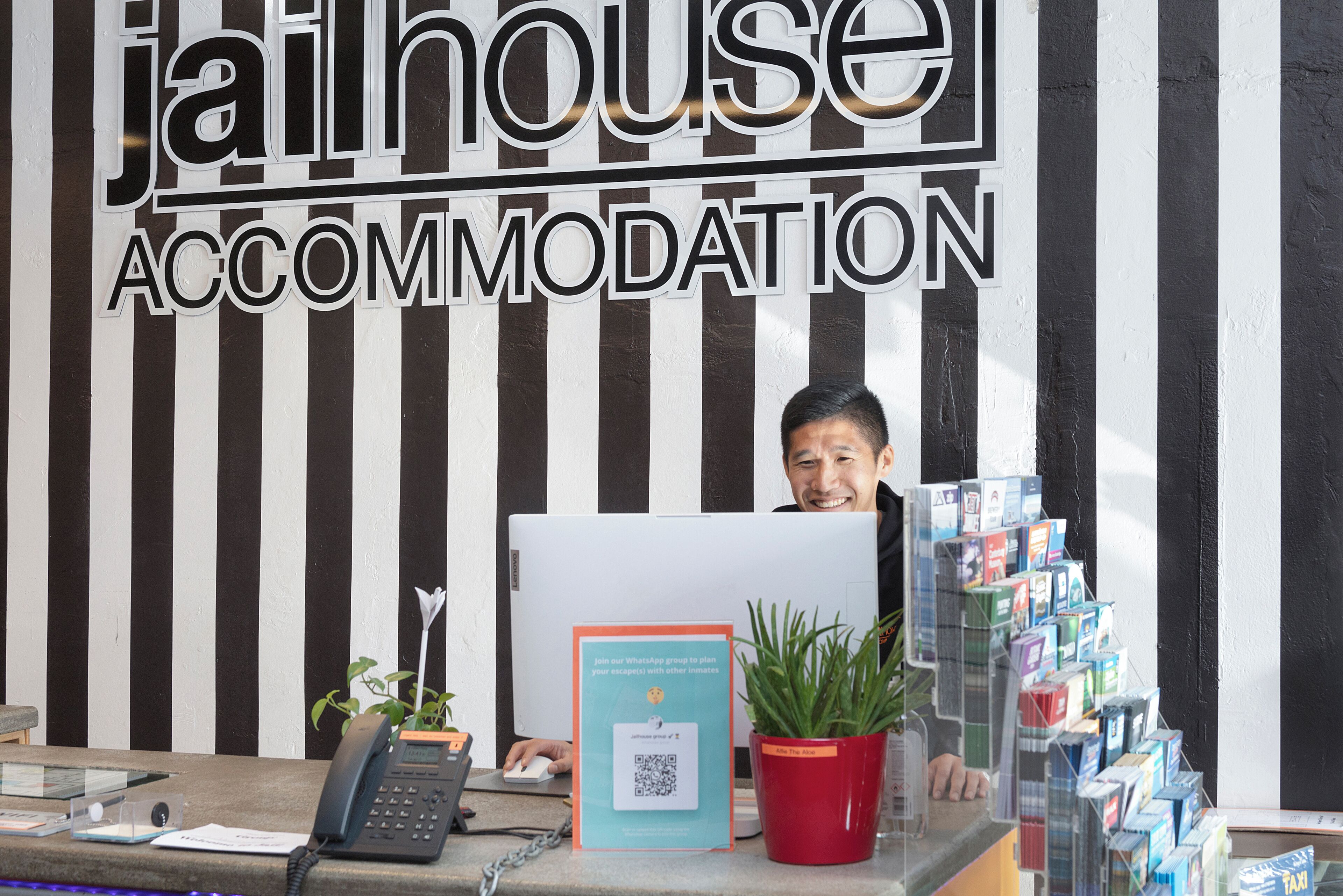 A reception desk with a computer, a phone, a QR code poster, a red potted plant, and a stack of brochures.
