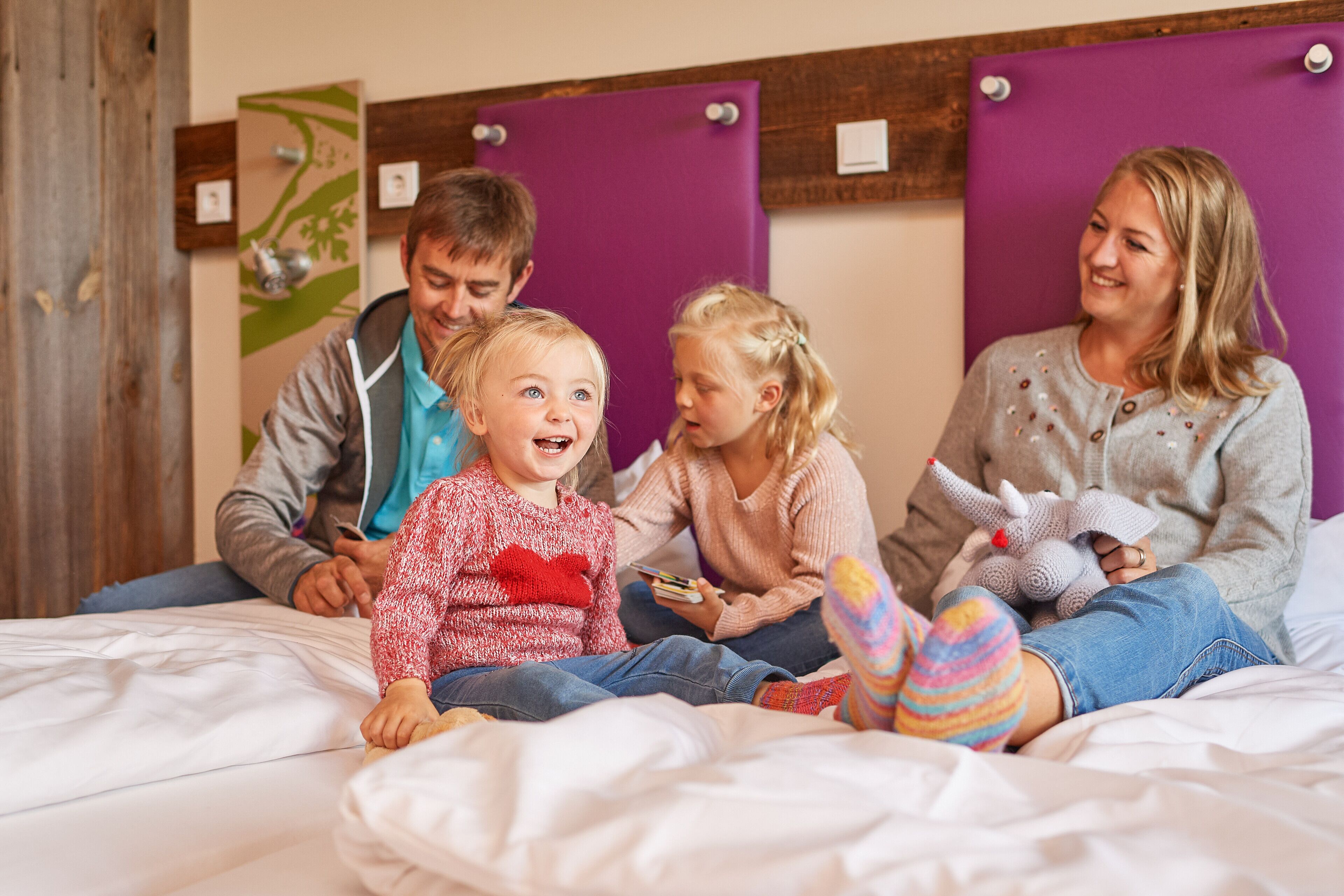 A family sitting on a bed with a purple headboard, a wooden wall panel, and a small plant decoration.