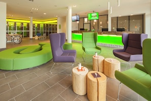 A modern reception area with green and purple seating, wooden tables, and a TV screen displaying a program. - Explorer Hotel Oberstdorf (Fischen im Allgaeu)