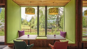 Espaço de refeições com duas mesas e cadeiras, amplas janelas com vista para vegetação e montanhas, e luminárias pendentes.