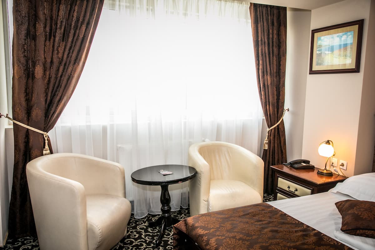 A hotel room with a bed, two armchairs, a small table, a nightstand with a lamp, and a window with curtains.
