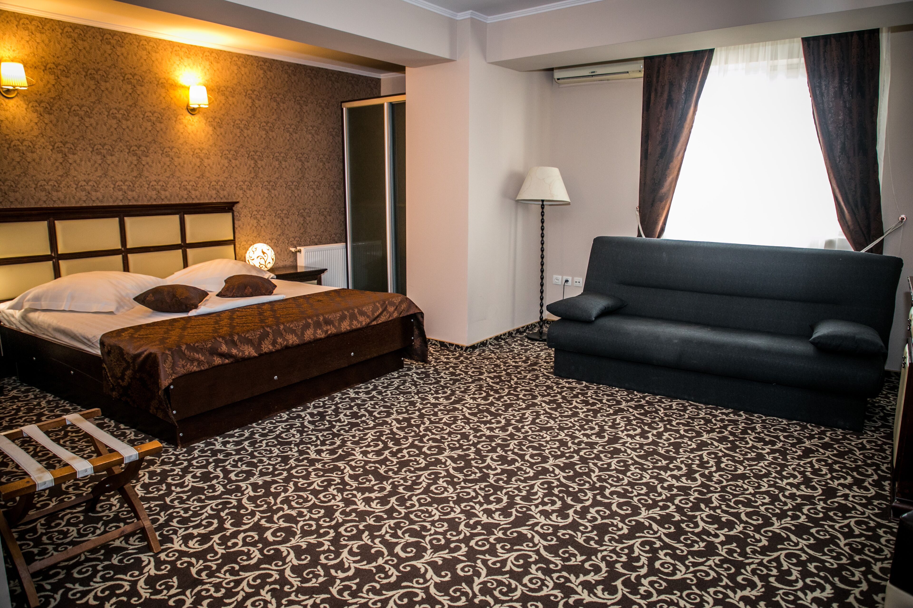 Une chambre d’hôtel avec un lit, un canapé, une moquette à motifs et une lampe fixée au mur.