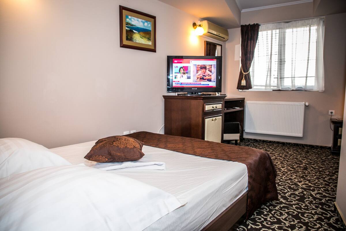 A hotel room with a bed, a television, a window with curtains, and a radiator.