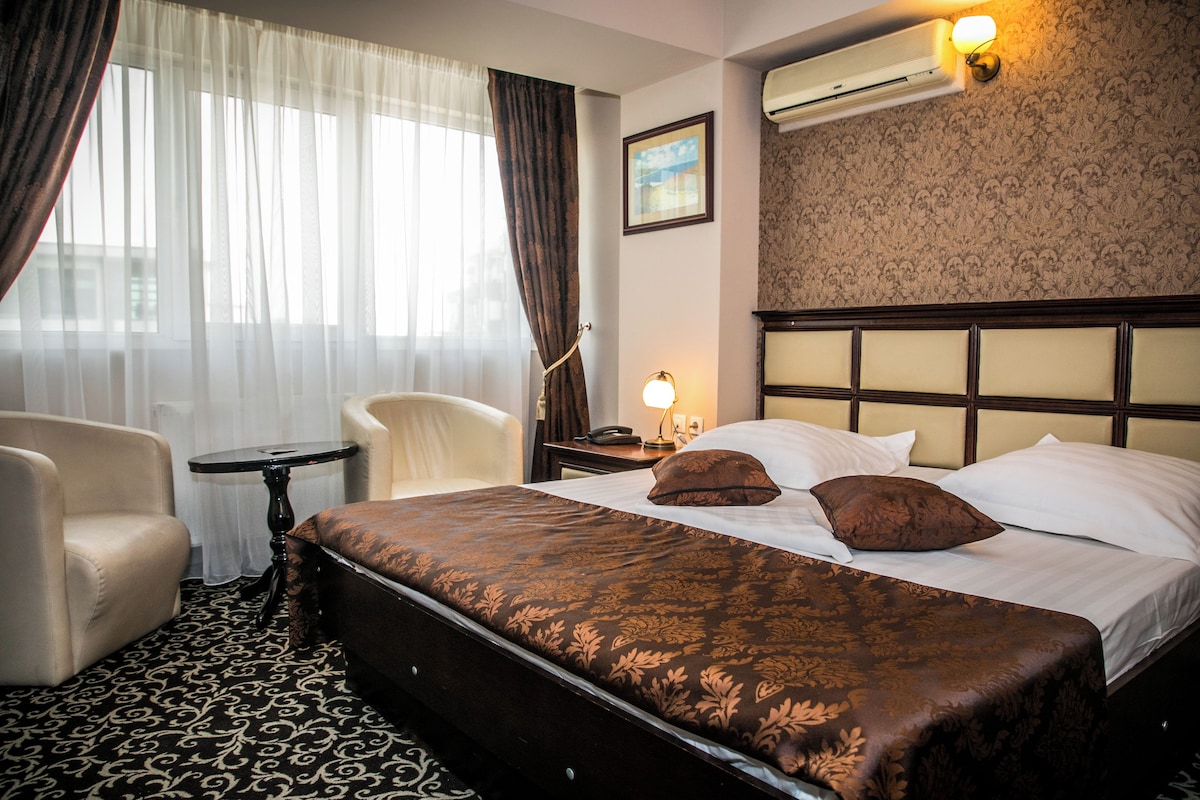 A hotel room with a large bed, a chair, a small table, and an air conditioning unit on the wall.