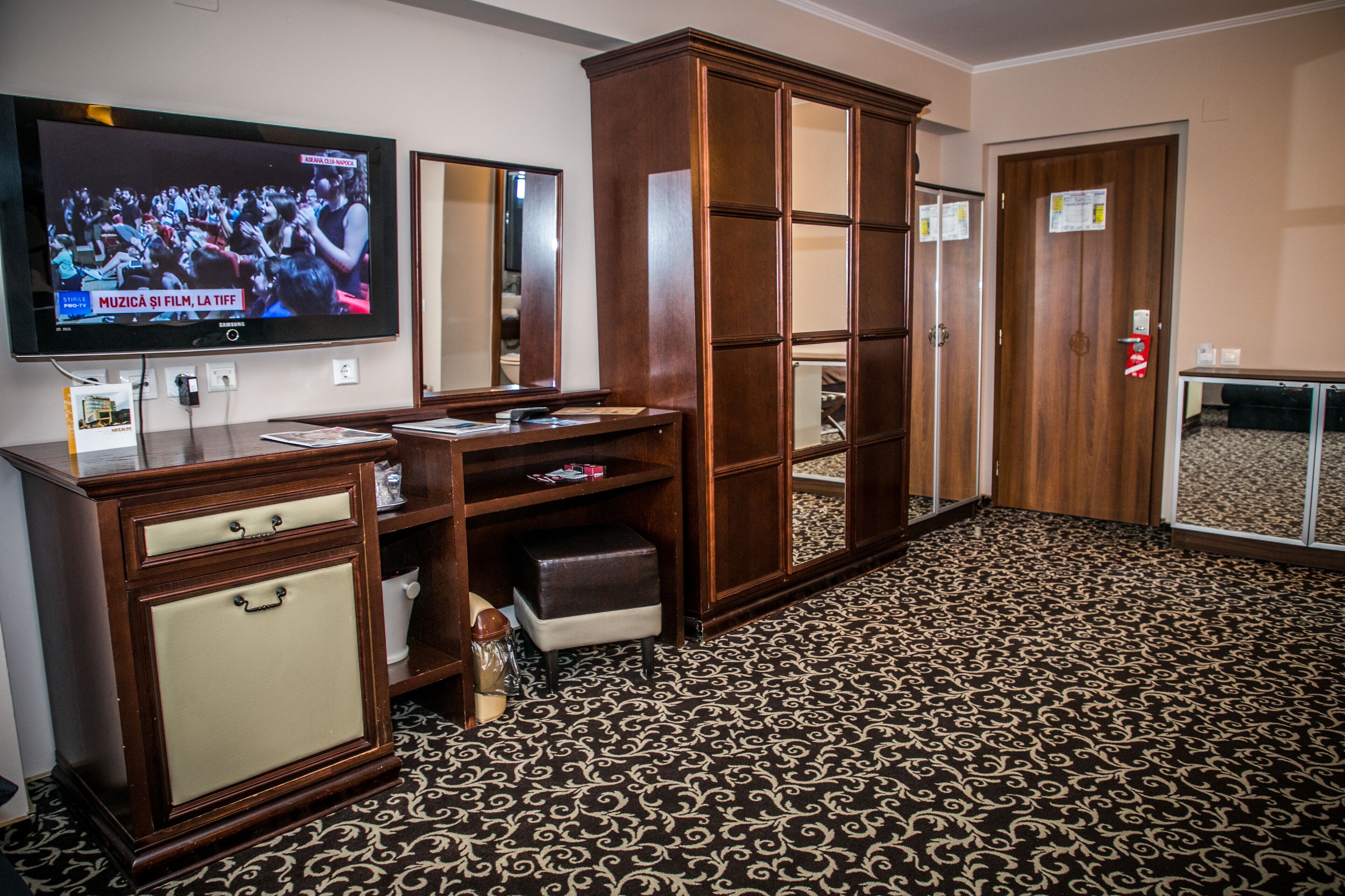 A hotel room with a large flat-screen TV, a wooden wardrobe, a mirror, and a patterned carpet.