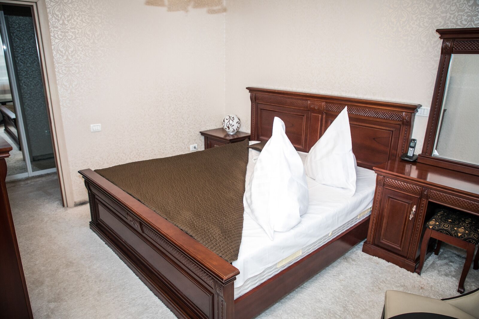 A neatly made bed with a wooden headboard, a bedside table with a vase, and a mirror on the wall.