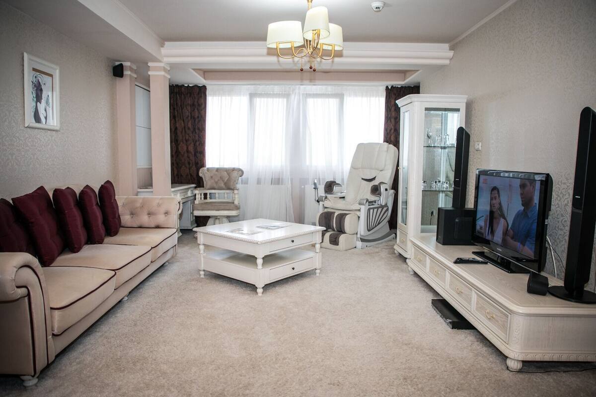 A living room with a beige sofa, a white coffee table, a massage chair, a TV, and a cabinet.