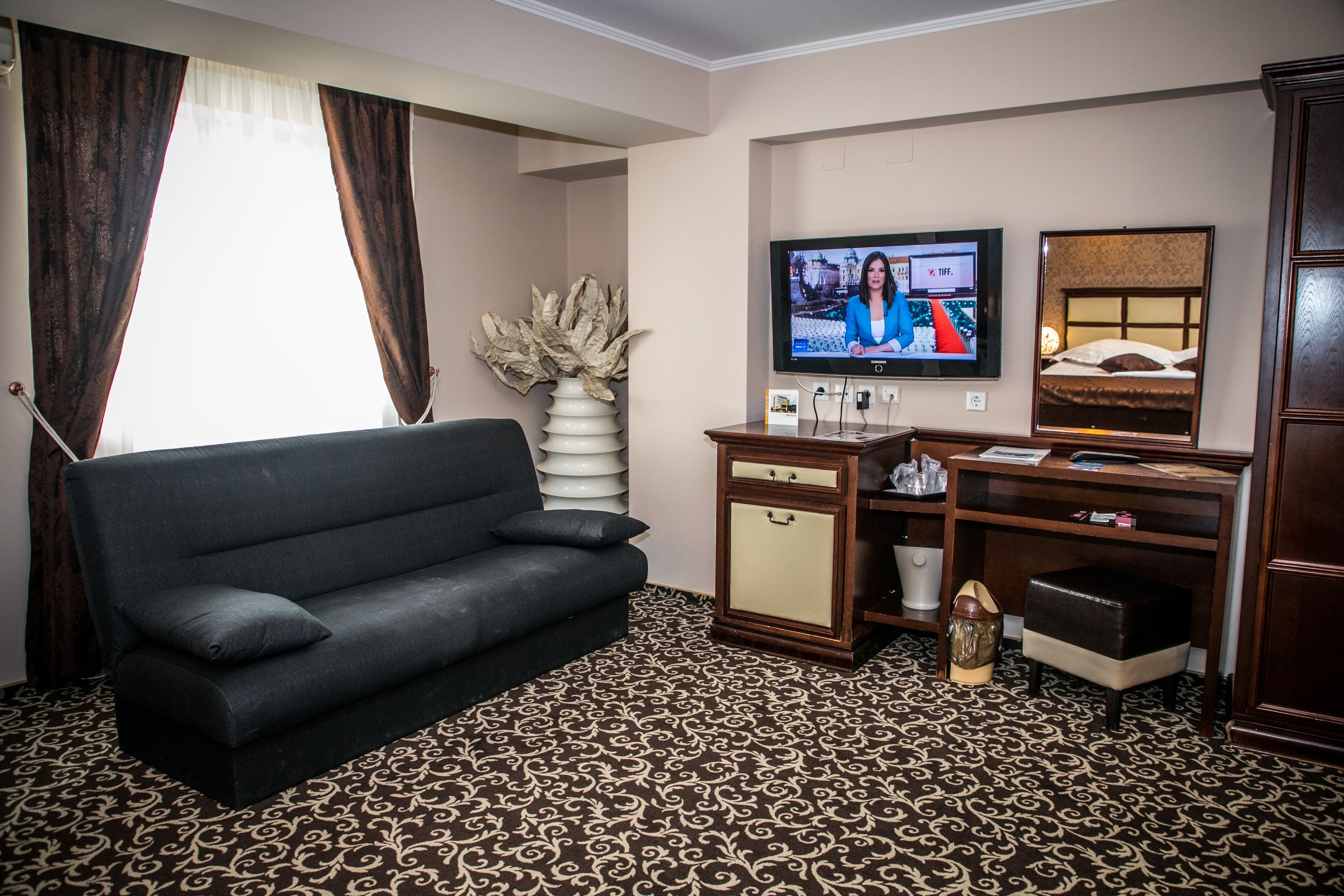 Habitación de hotel con sofá negro, televisor, escritorio de madera, armario y alfombra estampada.