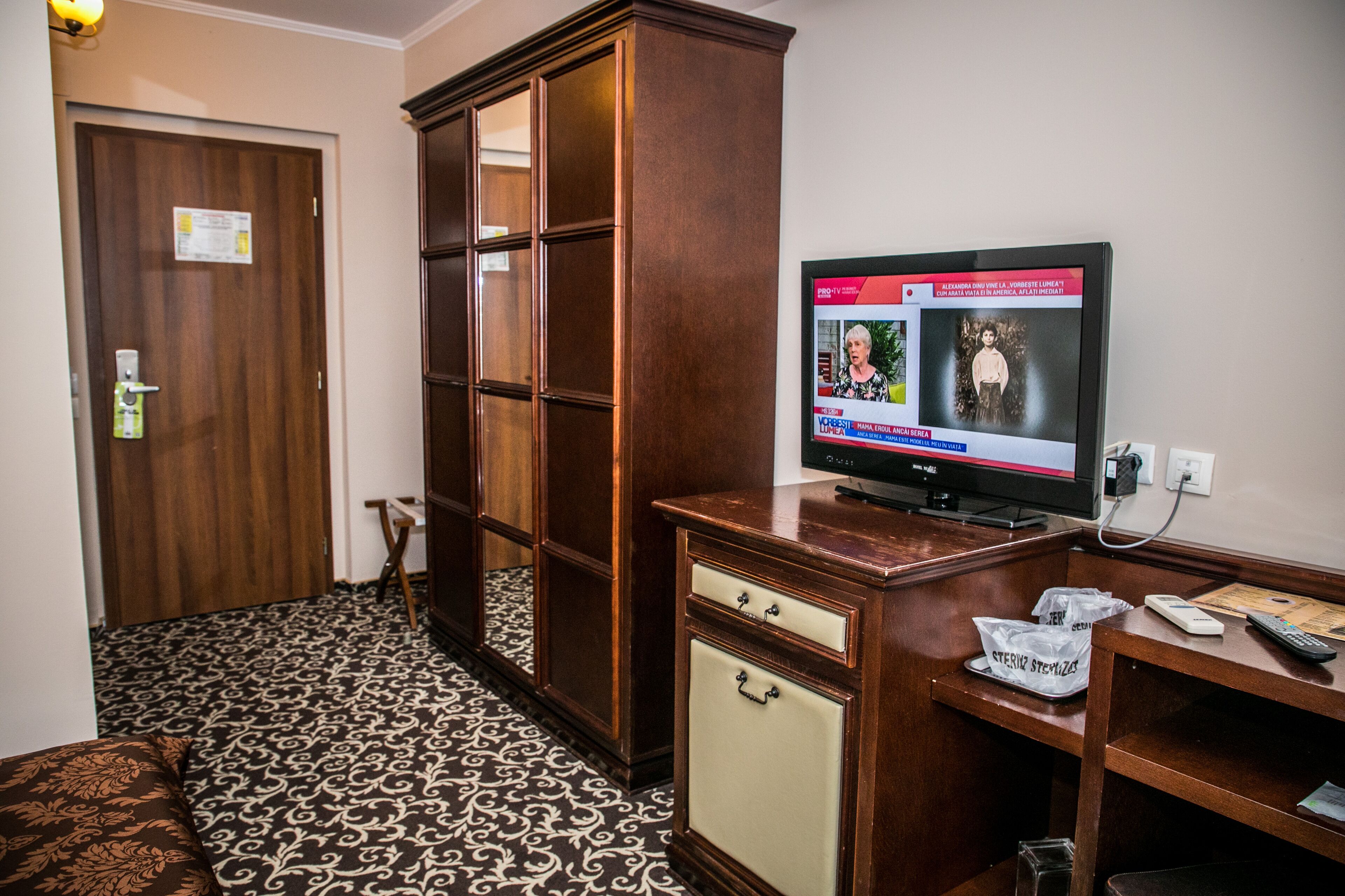 A hotel room with a wooden cabinet, a television, a desk with a telephone and a remote control, and a patterned floor.
