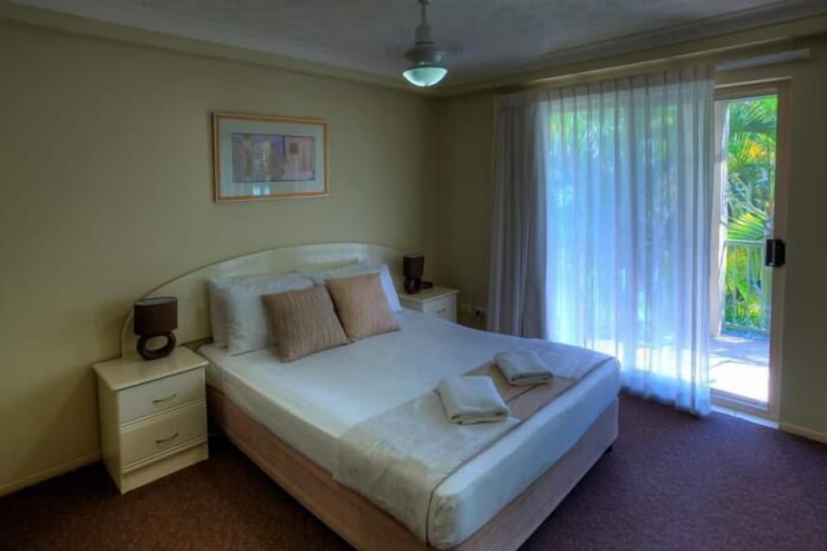 Una cama bien tendida con almohadas y una colcha, una mesita de noche con una lámpara y una ventana con cortinas.