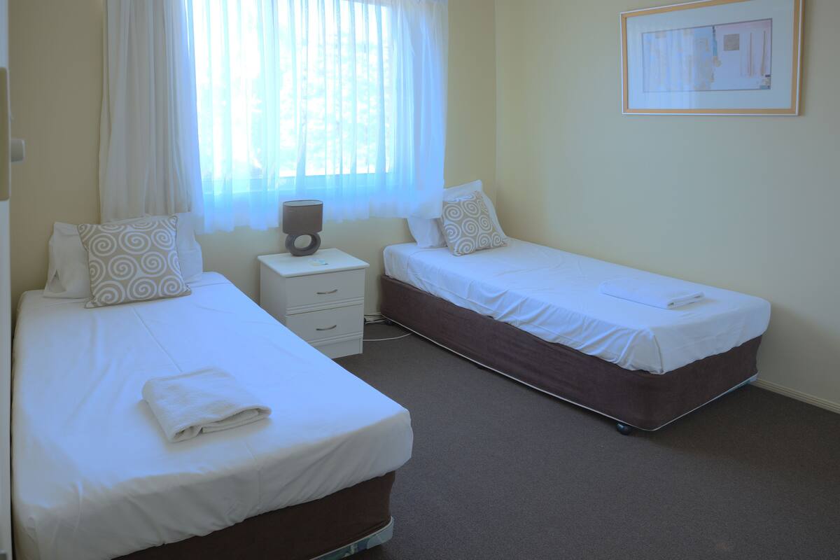 Habitación de hotel con dos camas individuales, una mesita de noche, una lámpara y un cuadro en la pared.