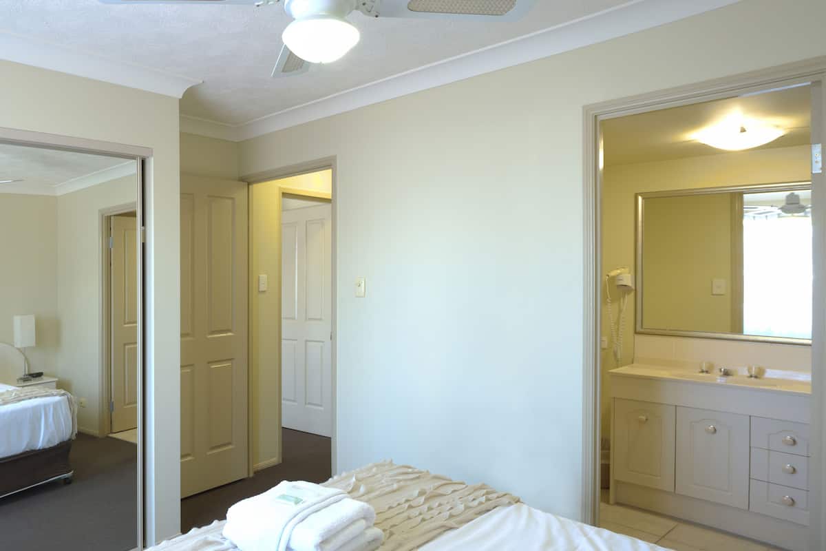 Habitación de hotel con cama, mesita de noche, espejo y baño con lavamanos y armarios.