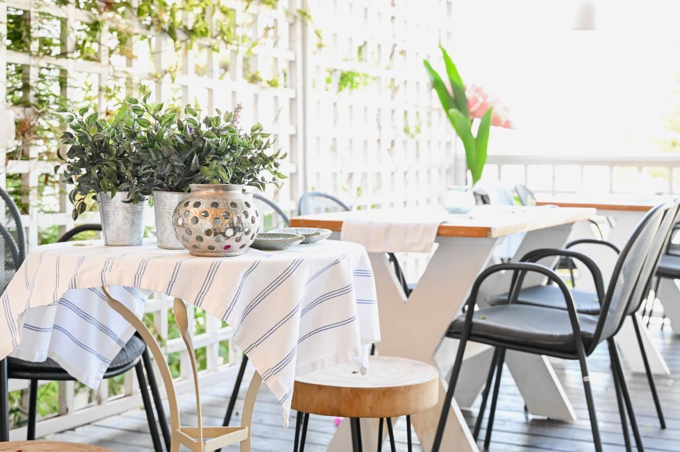 An outdoor dining area with tables and chairs, potted plants, and a white fence in the background.