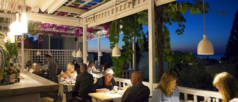 A restaurant with outdoor seating, white columns, hanging lights, and a view of the sky.